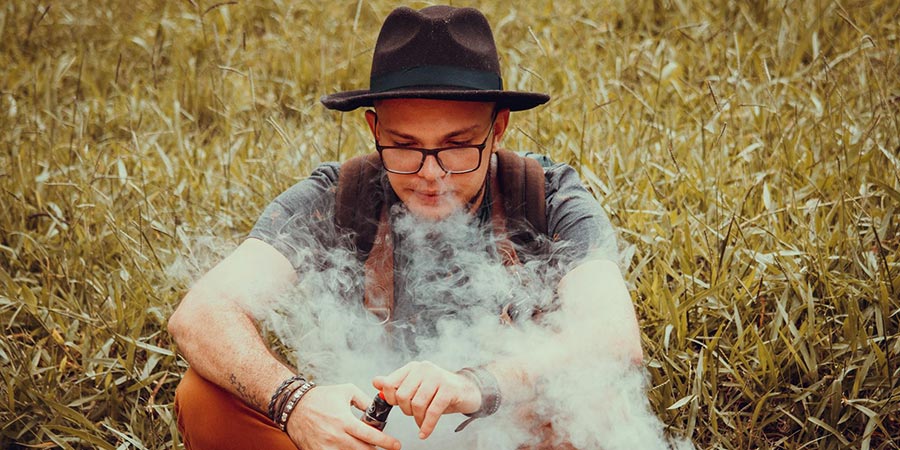 Man wearing glasses, gray shirt and brown cowboy hat sitting and vaping on a grass field